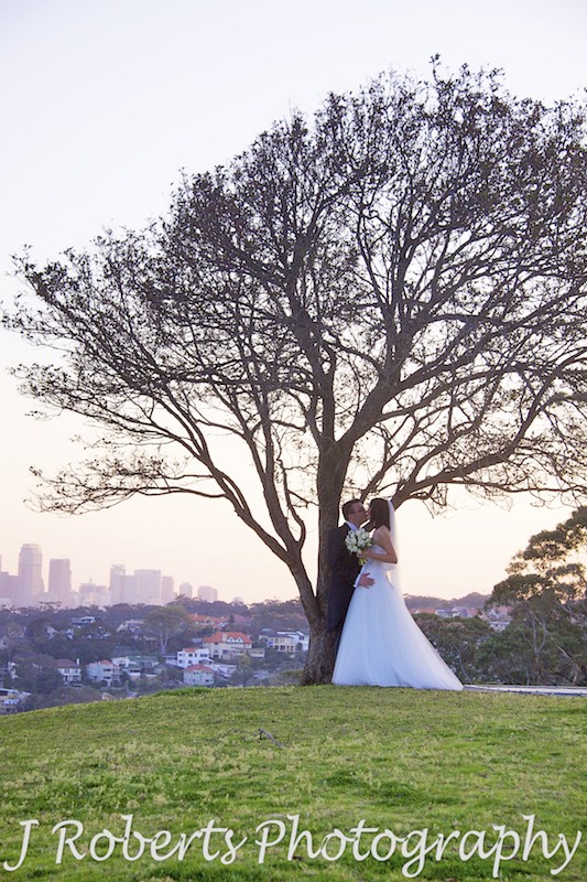 Bride and Groom kissing under tree with sunset and city skyline in the background - wedding photography sydney